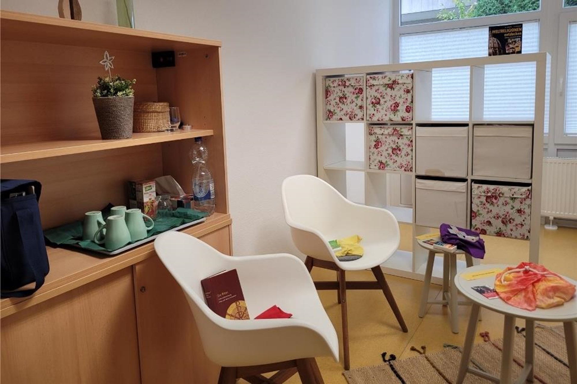 Ein heller, freundlicher Raum mit zwei weißen gepolsterten Schalensesseln um einen kleinen Beistelltisch und Regalen mit Büchern, Blumen und Kaffeegeschirr